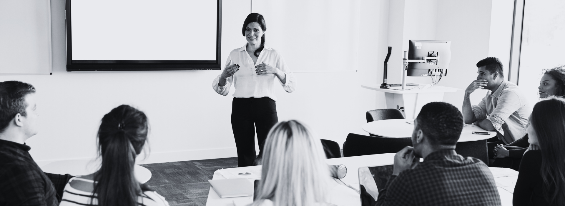 a woman in business attire addresses a room of people seated at desks